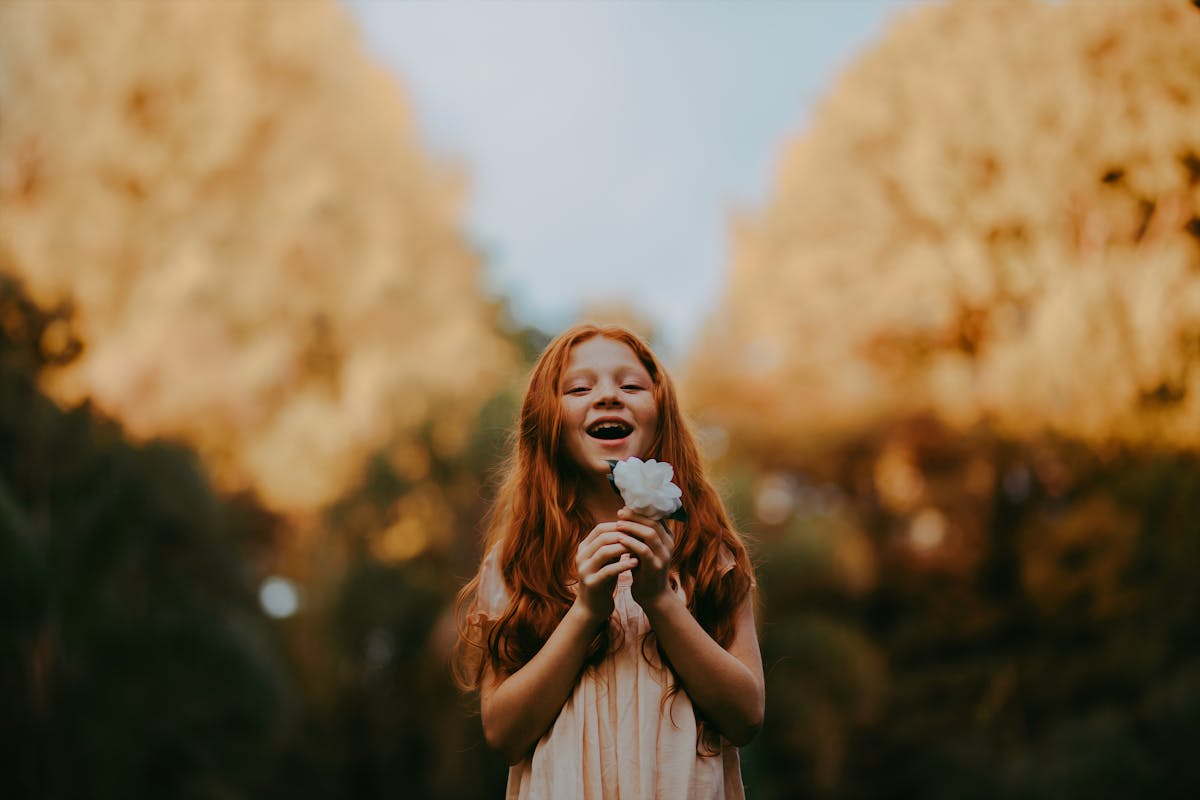 Smiling redheaded girl enjoys nature holding a flower outdoors during the day.