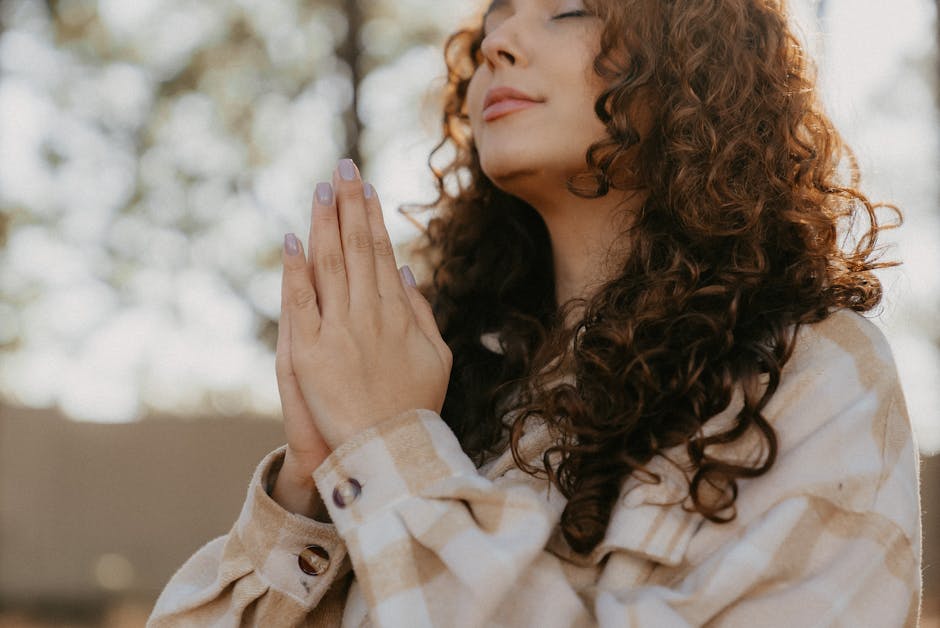 Serene woman with curly hair clasping hands in prayer outdoors. practice gratitude