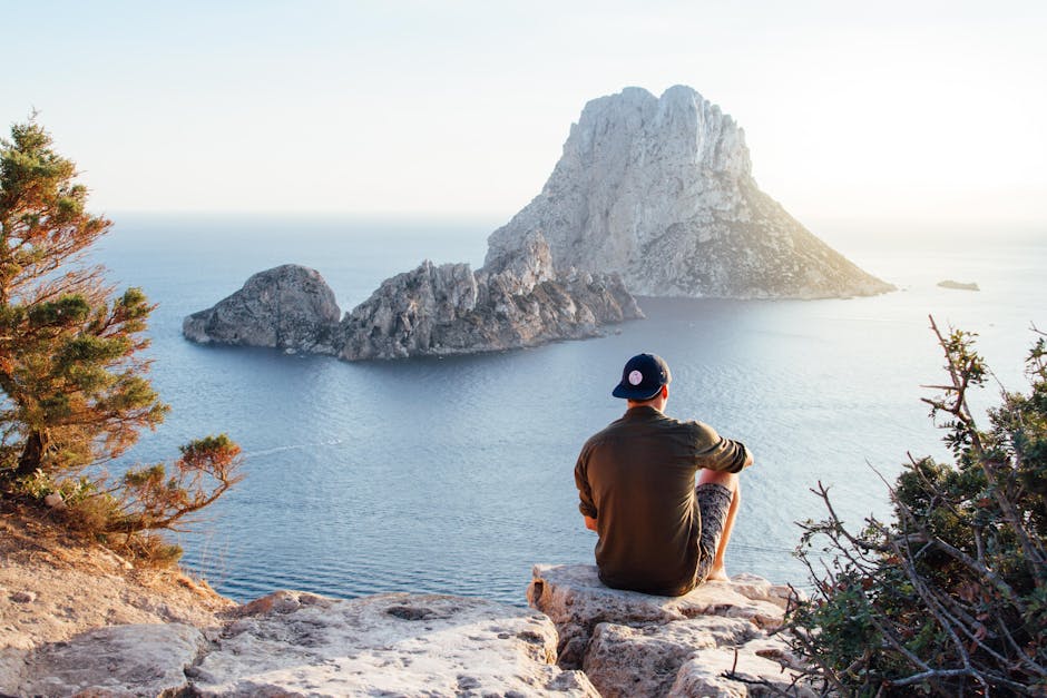 Man enjoys a scenic view of Es Vedrà at sunset from a cliff in San Juan Bautista, providing a perfect summer escape.