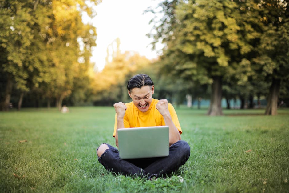 A young man in yellow celebrates success on his laptop while sitting in a sunlit park.