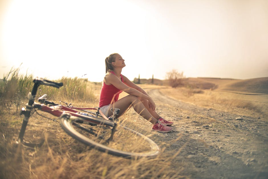 Full body of female in shorts and top sitting on roadside in rural field with bicycle near and enjoying fresh air with eyes closed - slow living